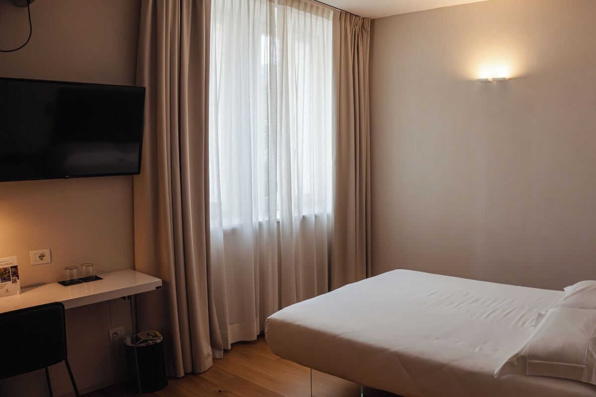A hotel room with a bed, a desk, a TV mounted on the wall, and a window with curtains.