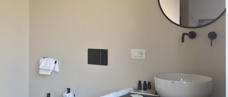 A modern bathroom with a large round mirror, a white sink, two bidets, a black shelf with towels, and a small black cabinet.