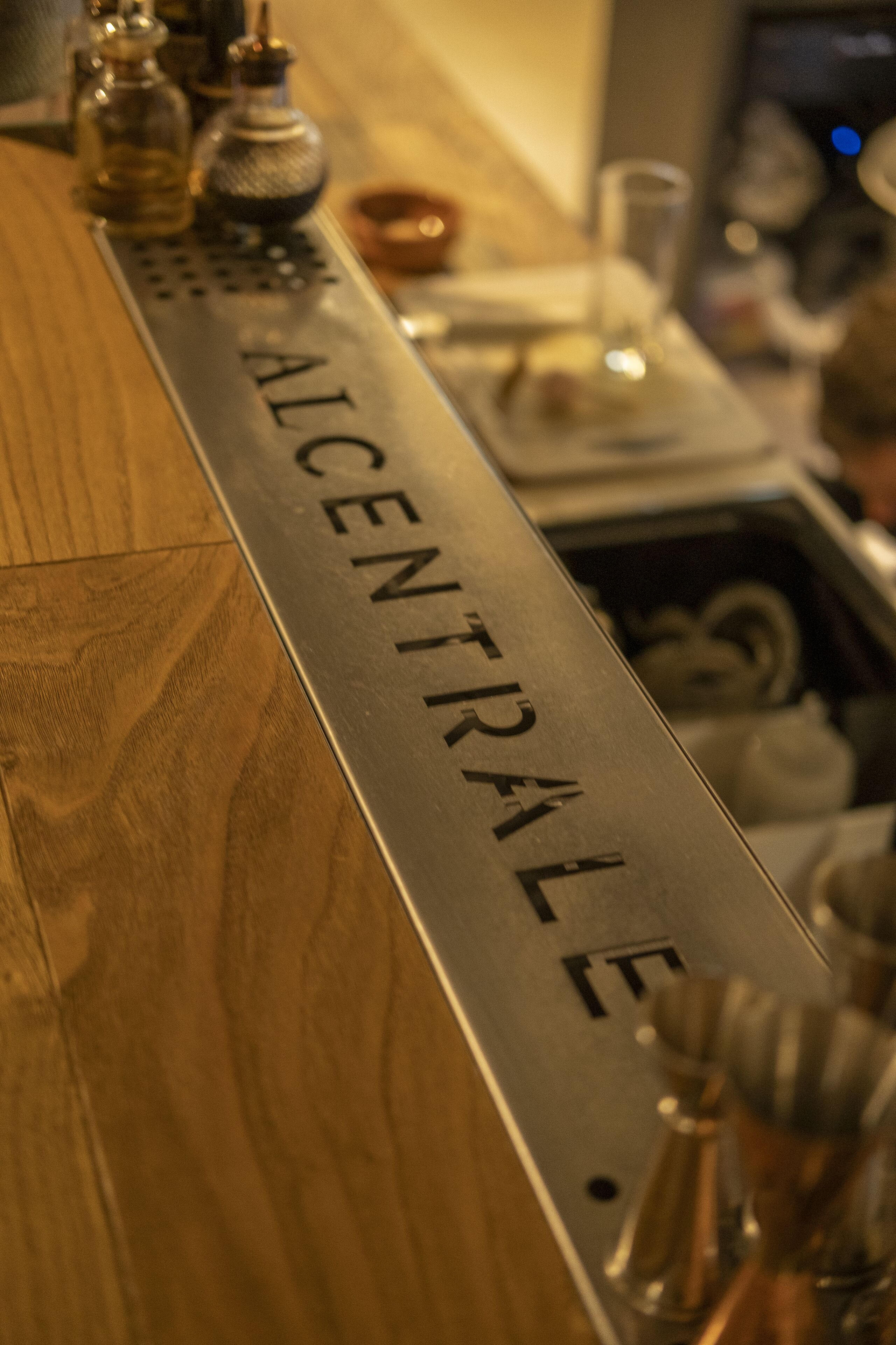 A bar counter with a metal sign reading "ALCENTRALE".