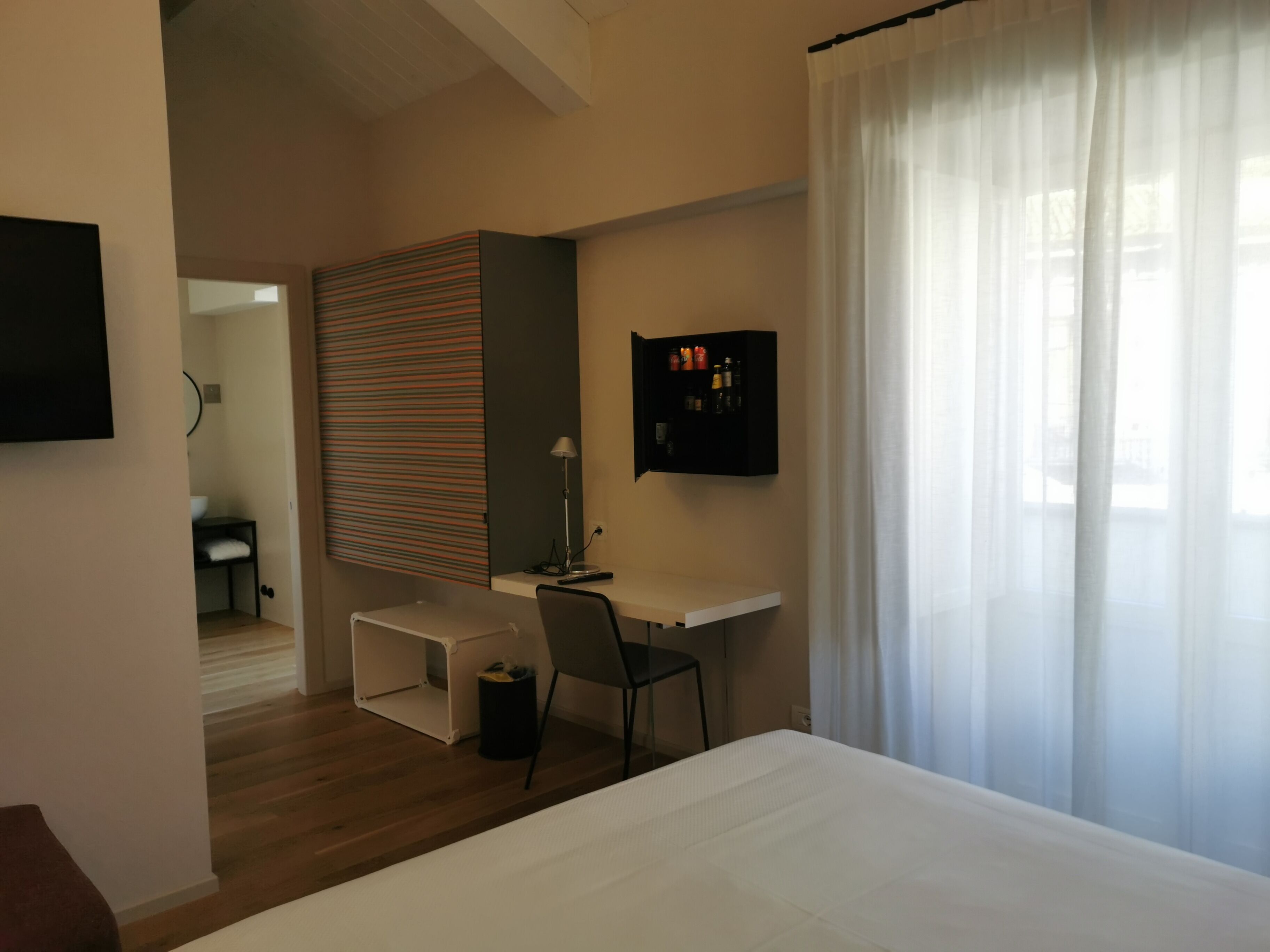 Ein modernes Schlafzimmer mit Bett, Schreibtisch und Stuhl. Ein Fernseher ist an der Wand befestigt und es gibt ein Fenster mit Vorhängen.