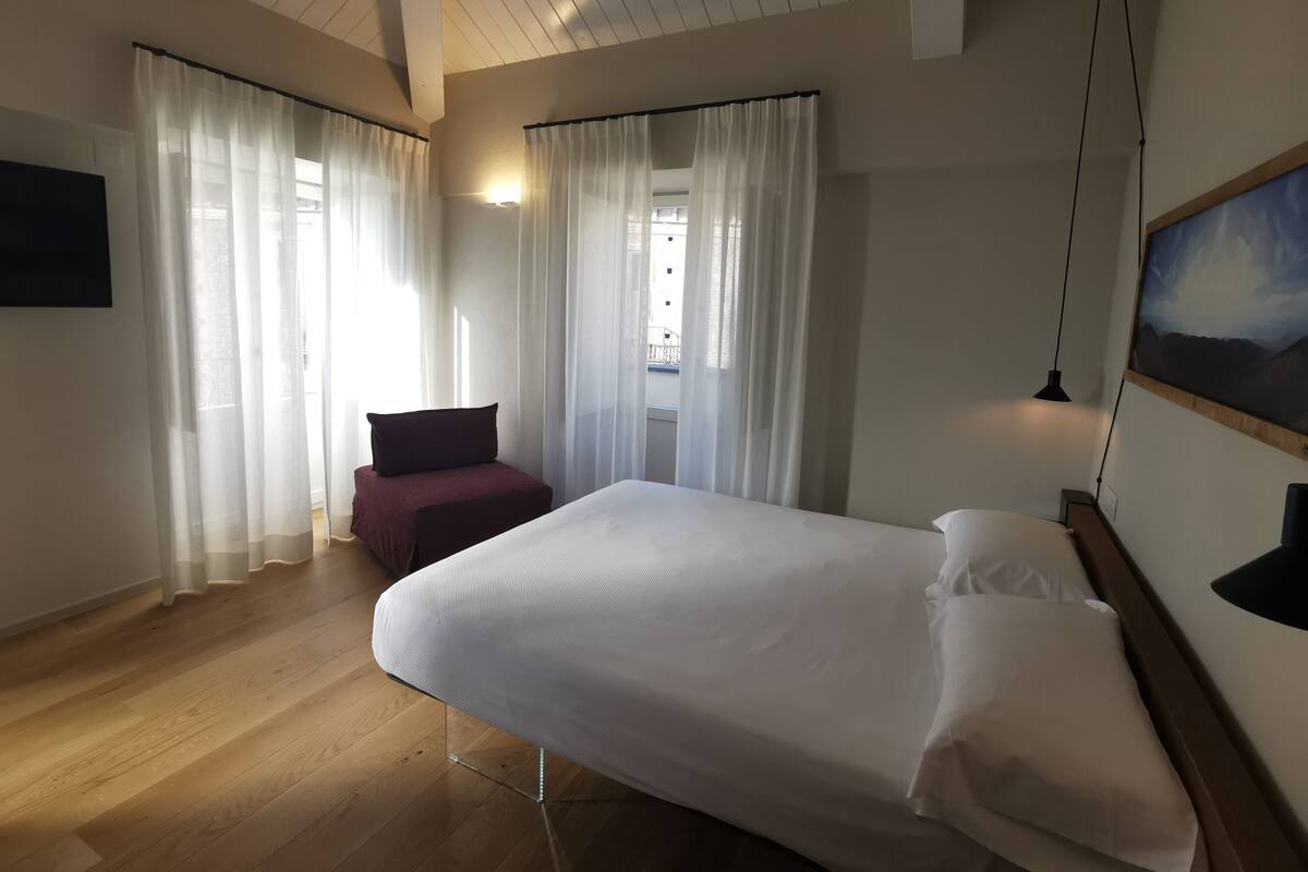 A bedroom with a bed, a chair, a painting, and a window with curtains.