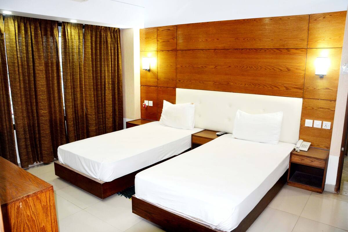 A hotel room with two single beds, wooden headboard, bedside tables with lamps, and a curtain.