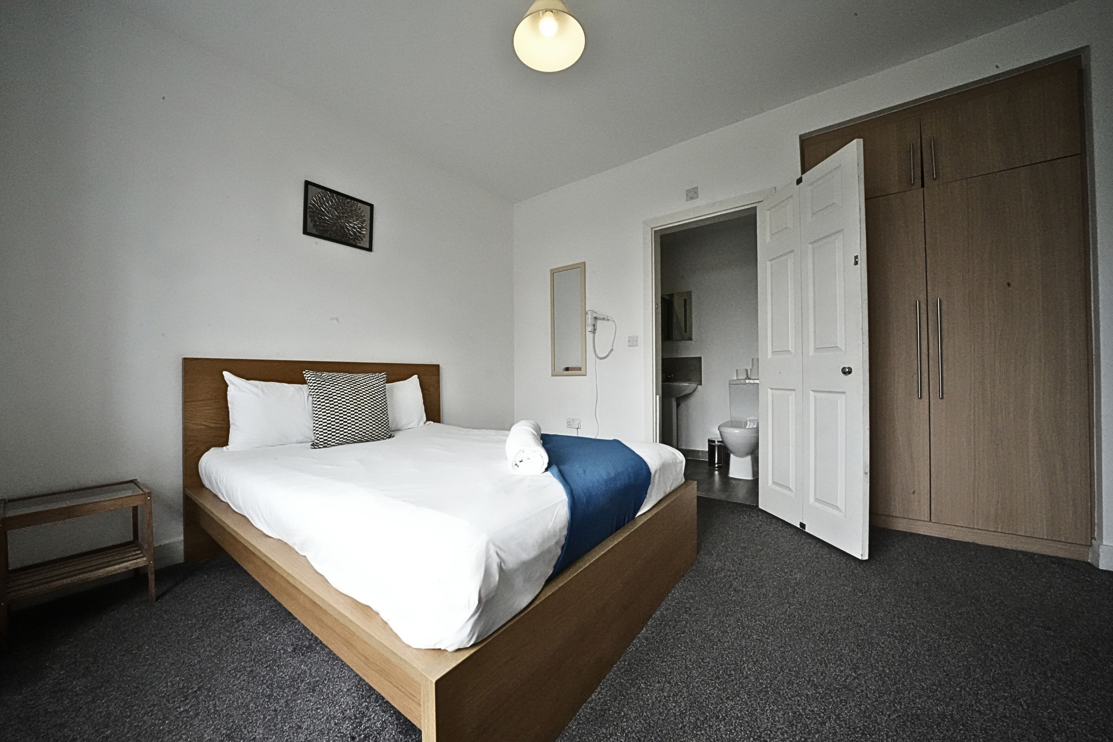 A bedroom with a wooden bed, white bedding, a small bedside table, a wardrobe, and an open door leading to a bathroom.