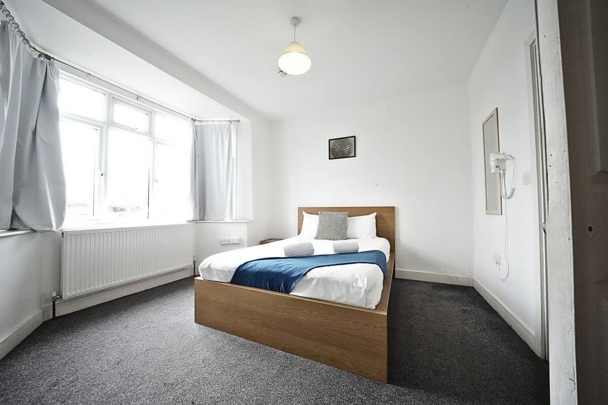 Ein Schlafzimmer mit einem hölzernen Bett, weißer Bettwäsche, einem Heizkörper, einem Fenster mit Vorhängen, einer Wandleuchte und einem Telefon.