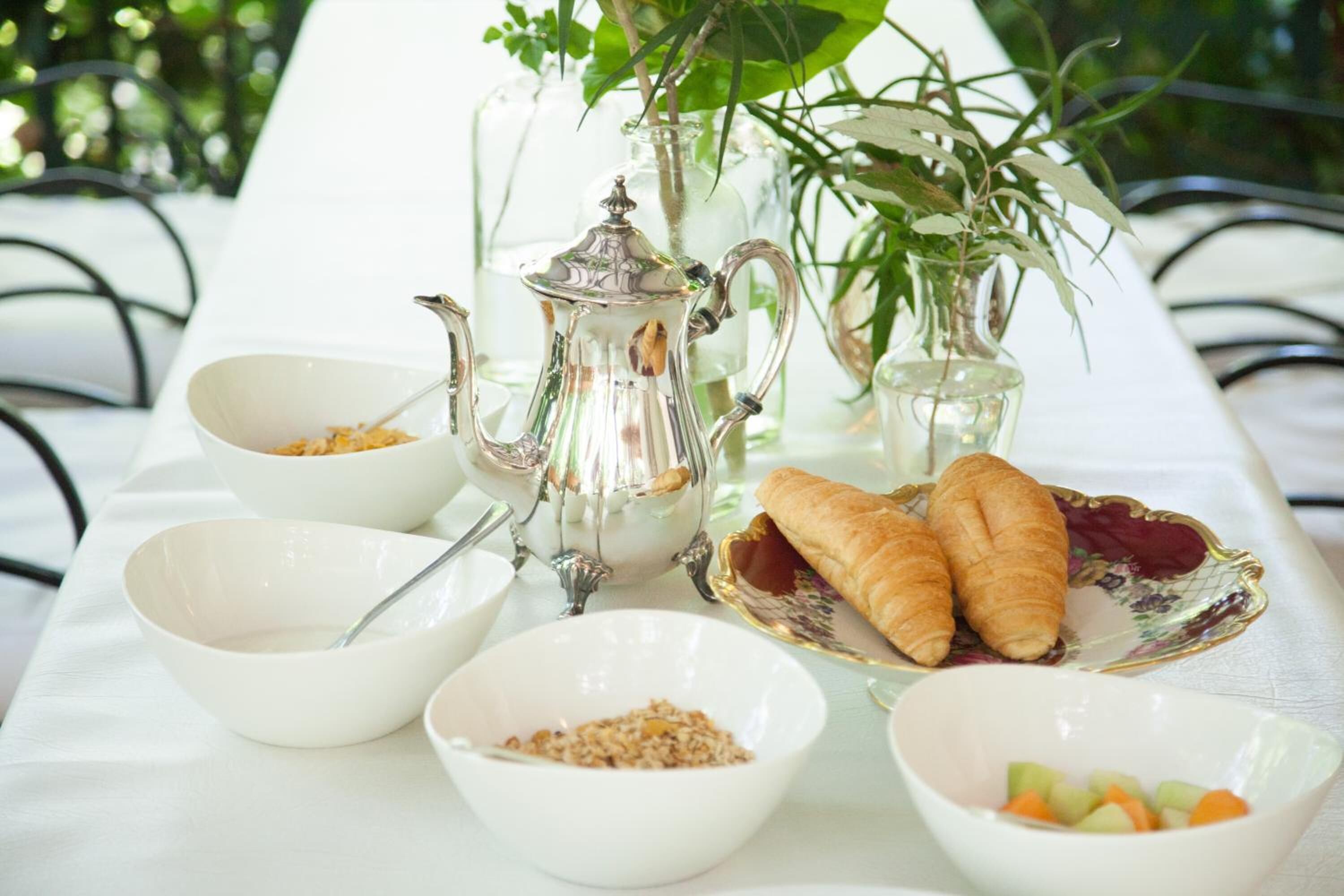 A table set with white bowls, a silver teapot, a glass pitcher, and croissants on a white tablecloth.