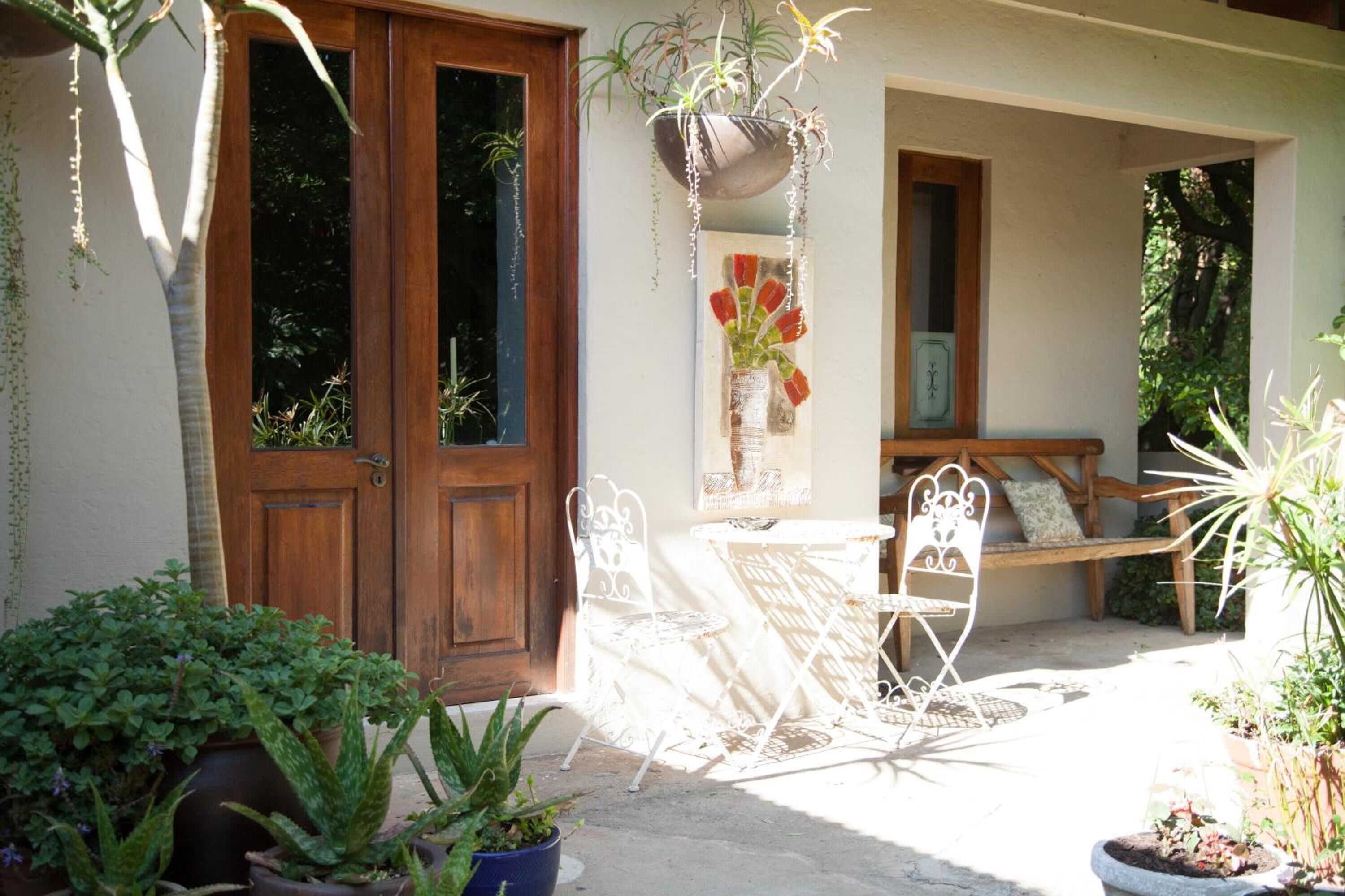 Une terrasse dotée d’une porte en bois, de chaises en métal blanc, d’une petite table et de divers plantes en pot.