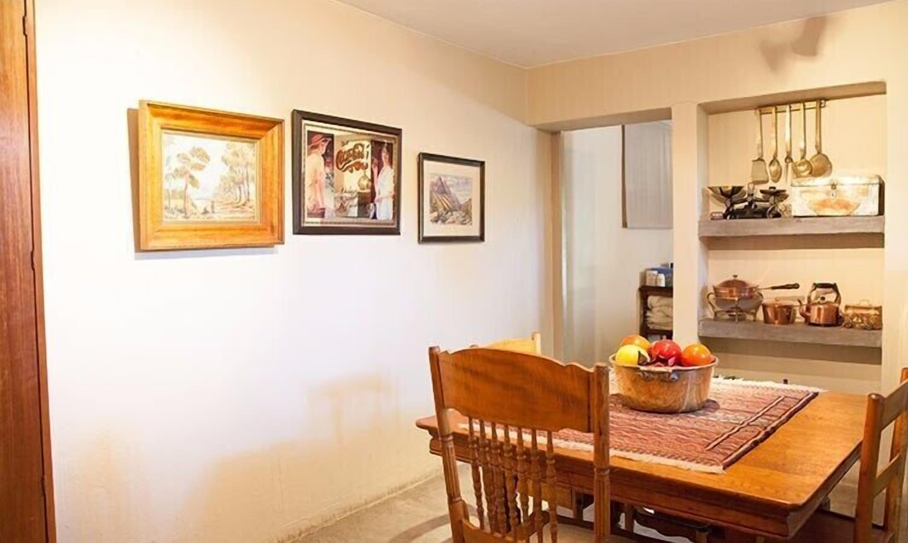 A dining area with a wooden table, chairs, and a fruit bowl. There are framed pictures on the wall and a shelf with kitchen utensils.