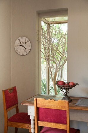 A dining area with a wooden table, two chairs, a wall clock, and a window with a view of a tree. - Little Forest Guest House (Randburg)