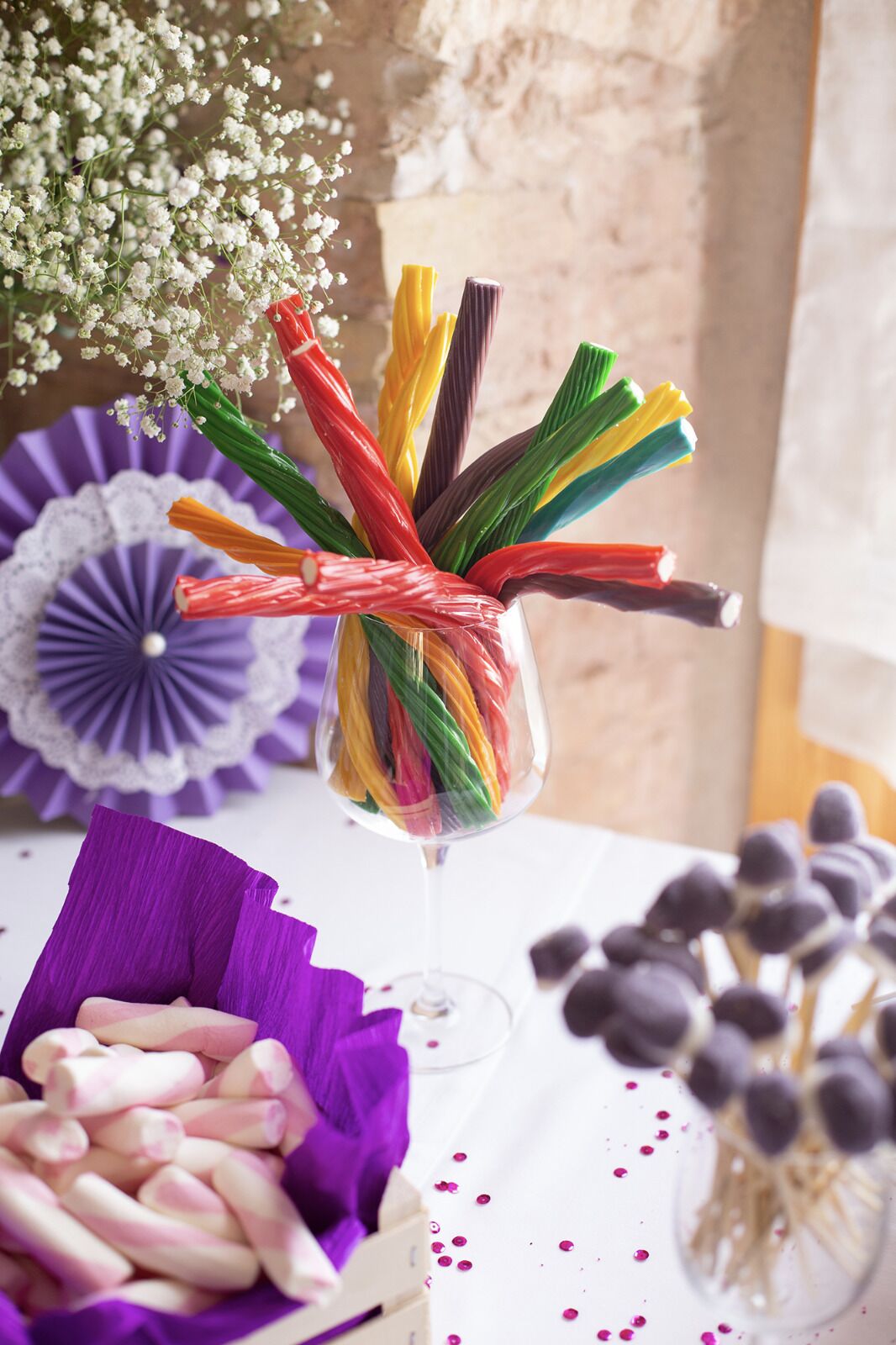 Une table sur laquelle se trouve un vase en verre contenant des crayons de couleur attachés par un ruban rouge, un éventail violet et un bol rempli d’objets roses et blancs.