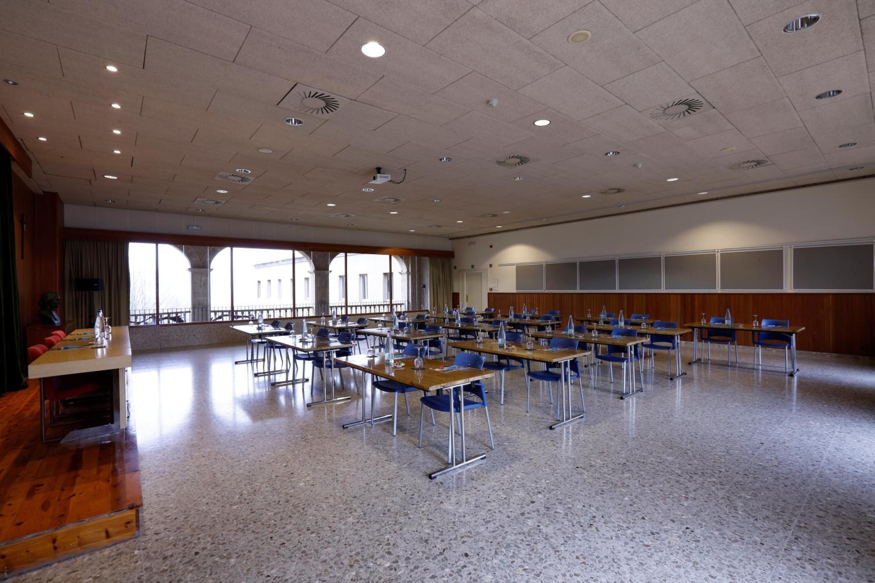 Un salón de conferencias con varias mesas y sillas, ventanas amplias y una zona de escenario.