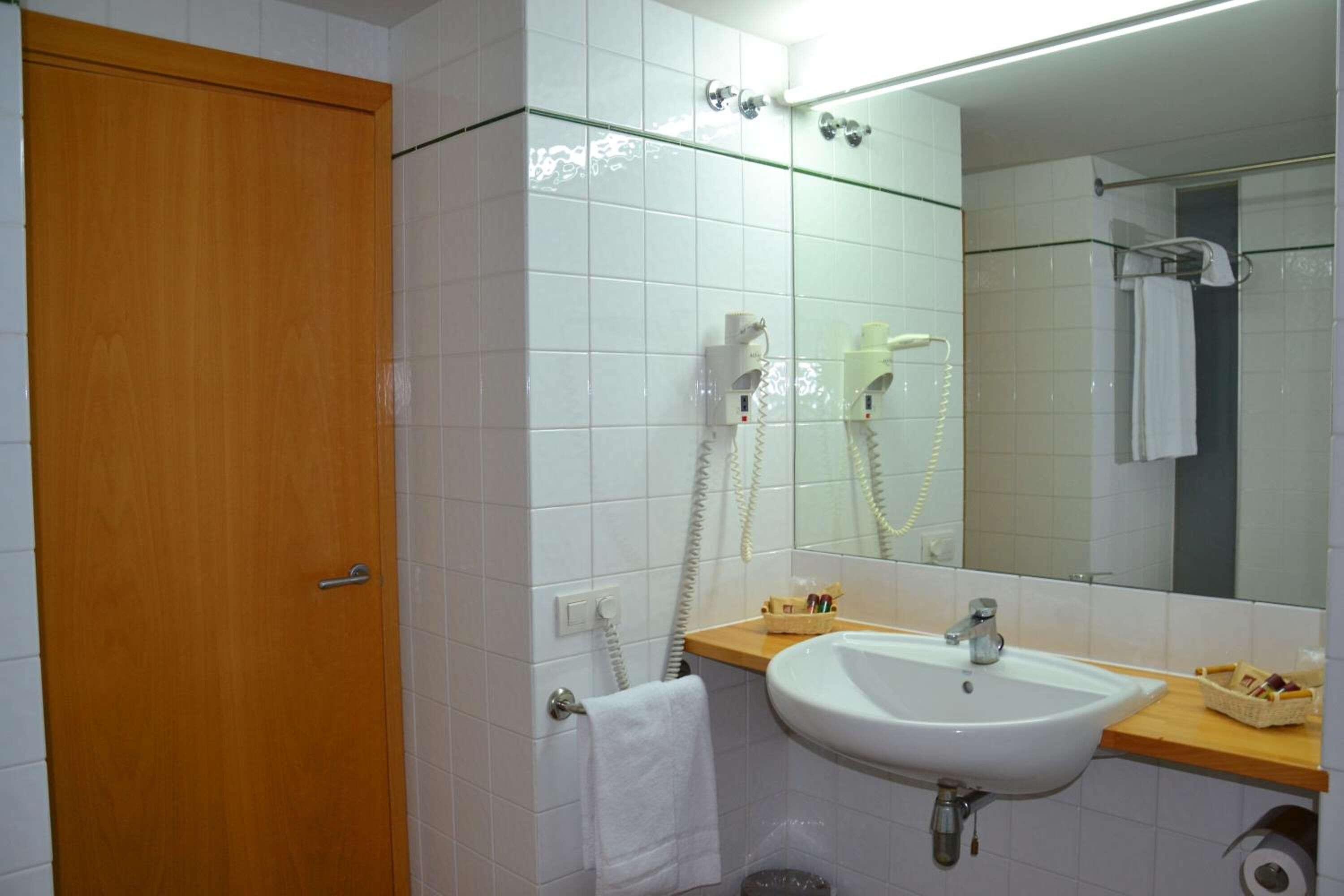 Un baño con puerta de madera, paredes con azulejos blancos, un lavabo blanco, un espejo, un perchero para toallas y un secador de pelo.