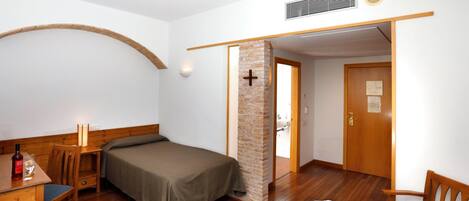 Una habitación pequeña y acogedora con una cama, un escritorio y una silla. Hay una puerta de madera y un pilar de ladrillo con una cruz.