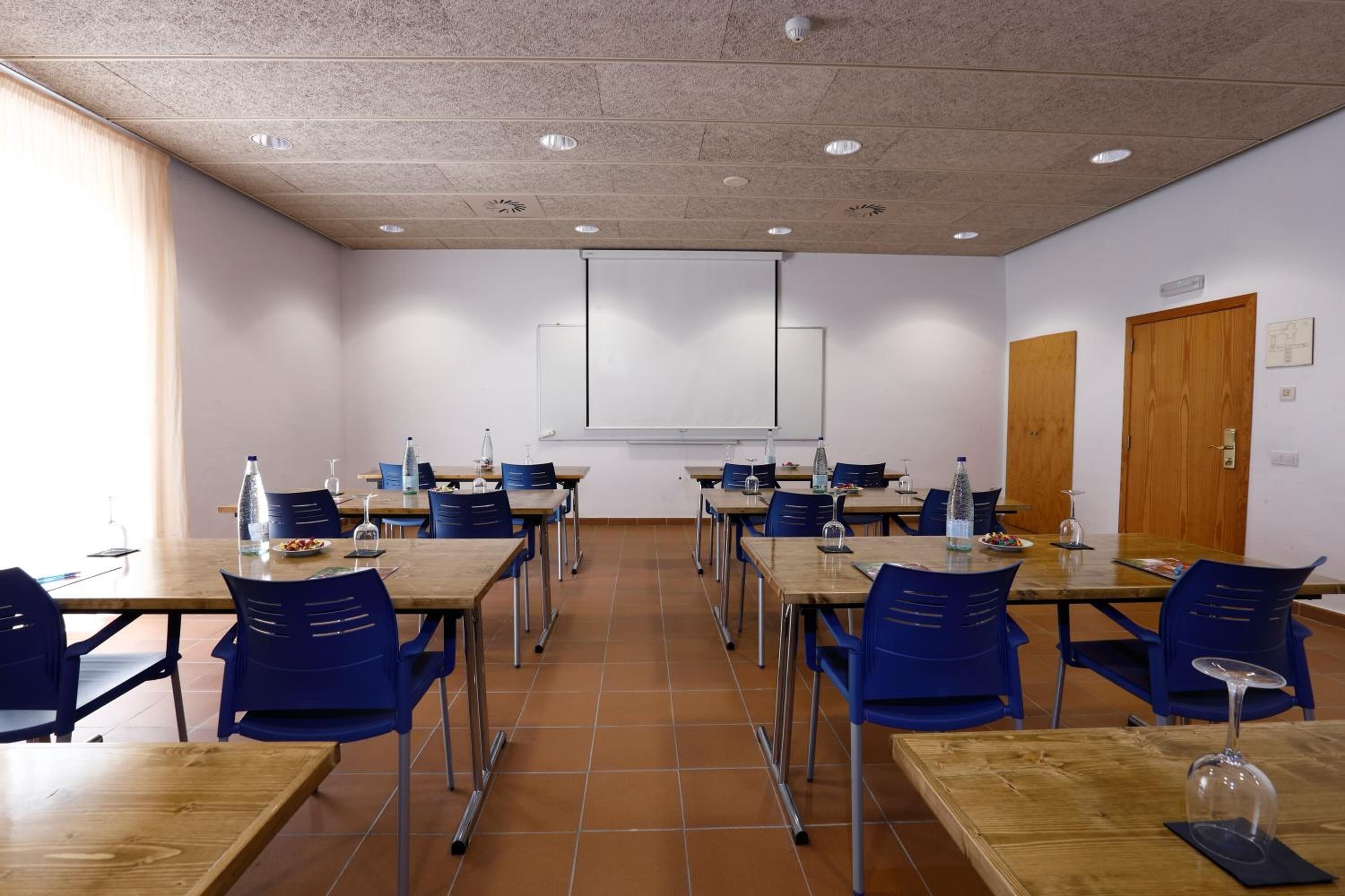 Un salón de conferencias con varias mesas y sillas, una pantalla de proyector y una puerta de madera.