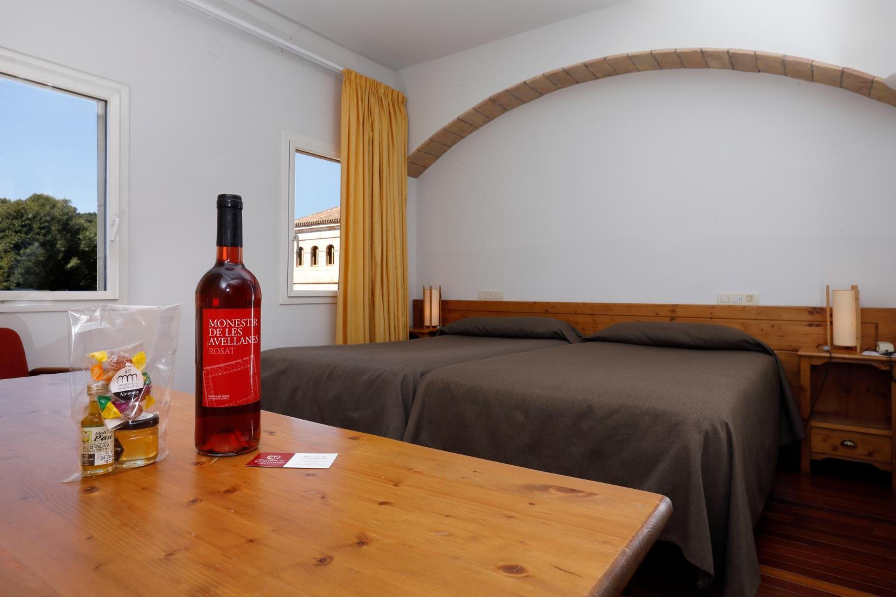 A hotel room with a bed, a wooden table with a bottle of rosé wine and a small bottle of honey, a window with a view of a building, and a curved brick arch above the bed.