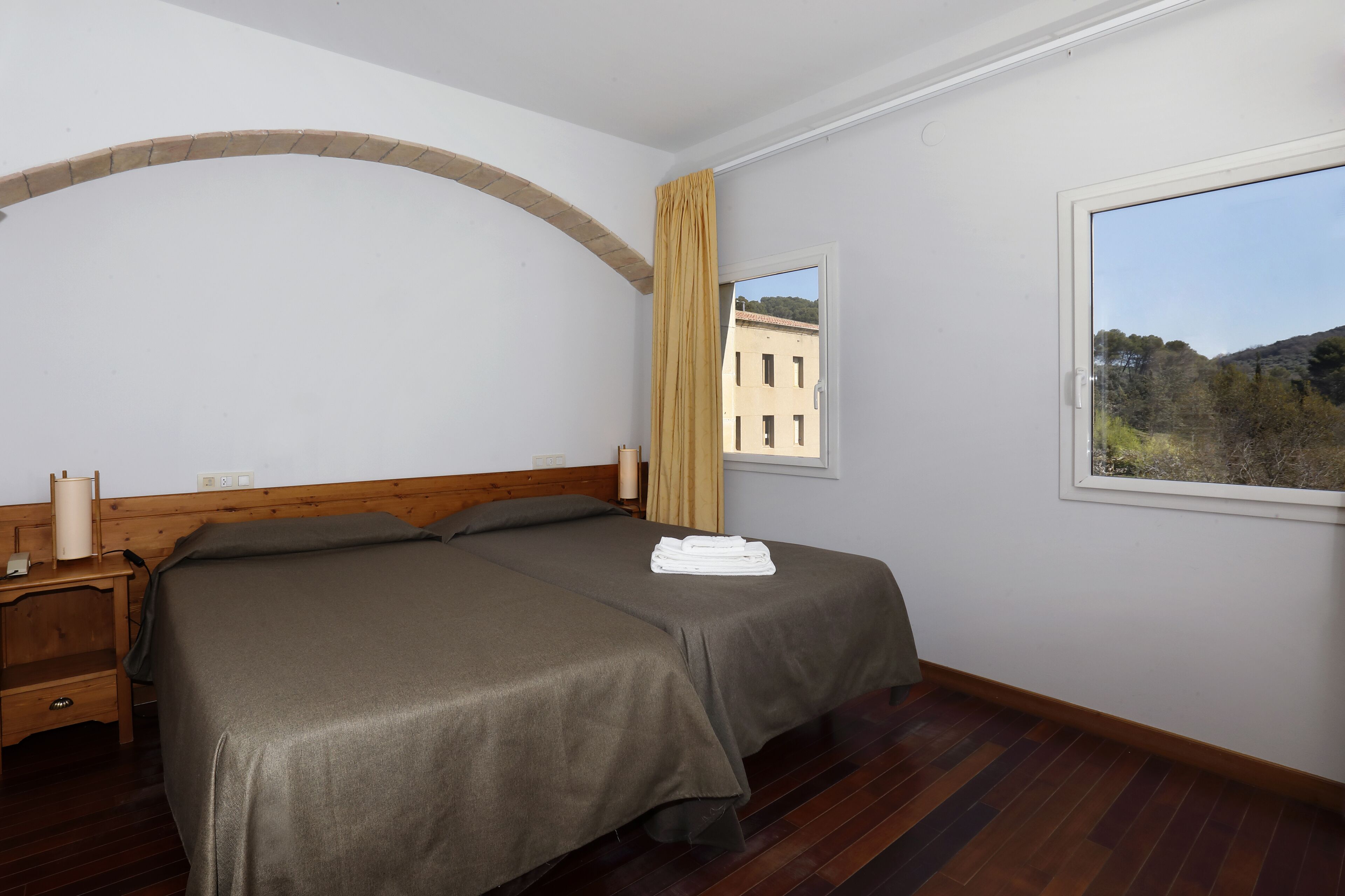 Un dormitorio con cama doble, cabecera de madera, una mesita de noche, una ventana con vistas a edificios y árboles, y una cortina.