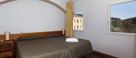 A bedroom with a double bed, wooden headboard, a nightstand, a window with a view of buildings and trees, and a curtain.