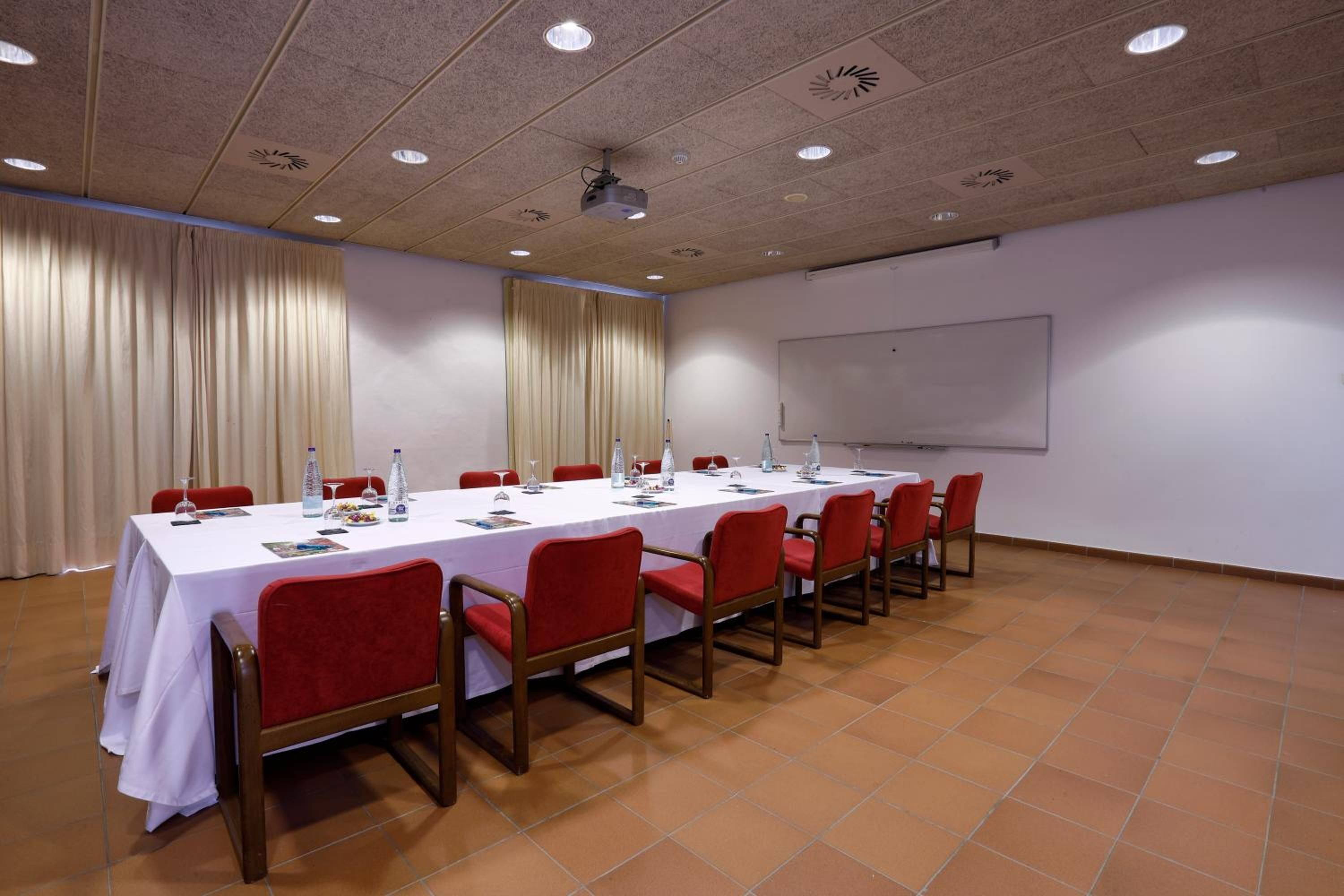 Un salón de conferencias con una larga mesa, sillas rojas, un proyector y una pizarra.
