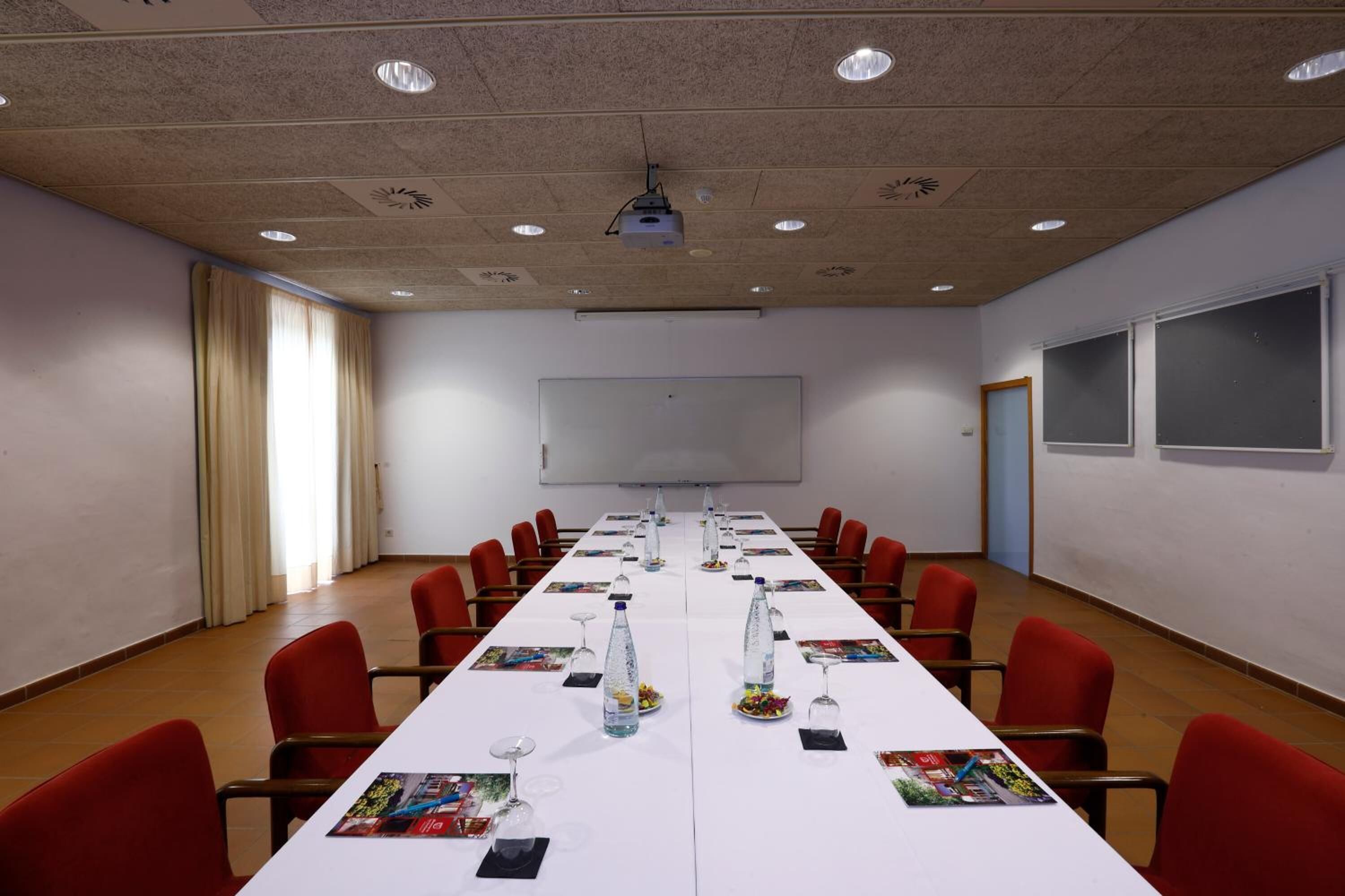 Ein Konferenzraum mit langem Tisch, roten Stühlen, einem Projektor und einer Whiteboard-Tafel.