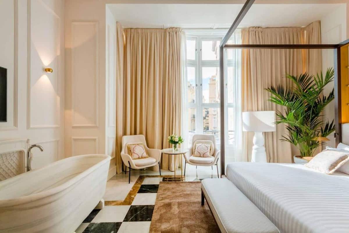 A hotel room with a large bed, a sitting area with two chairs, a small table with a vase, a bathtub, and a large window with curtains.