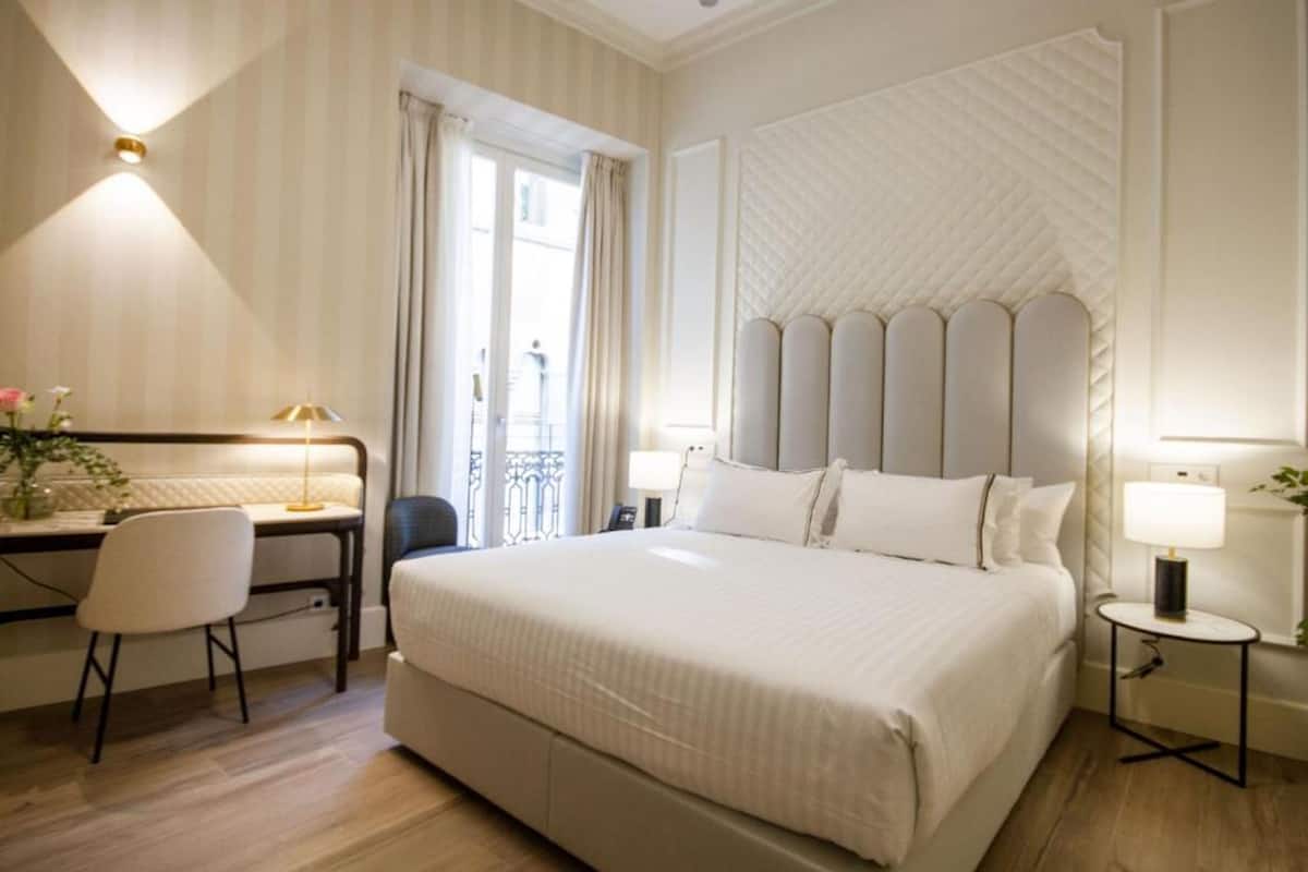 A hotel room with a large bed, a desk with a chair, a small table with a lamp, and a window with curtains.