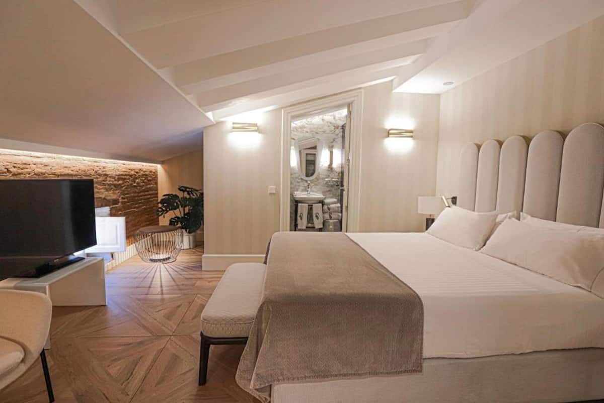 A modern hotel room with a large bed, a flat-screen TV, a seating area with a bench, and a stone accent wall.