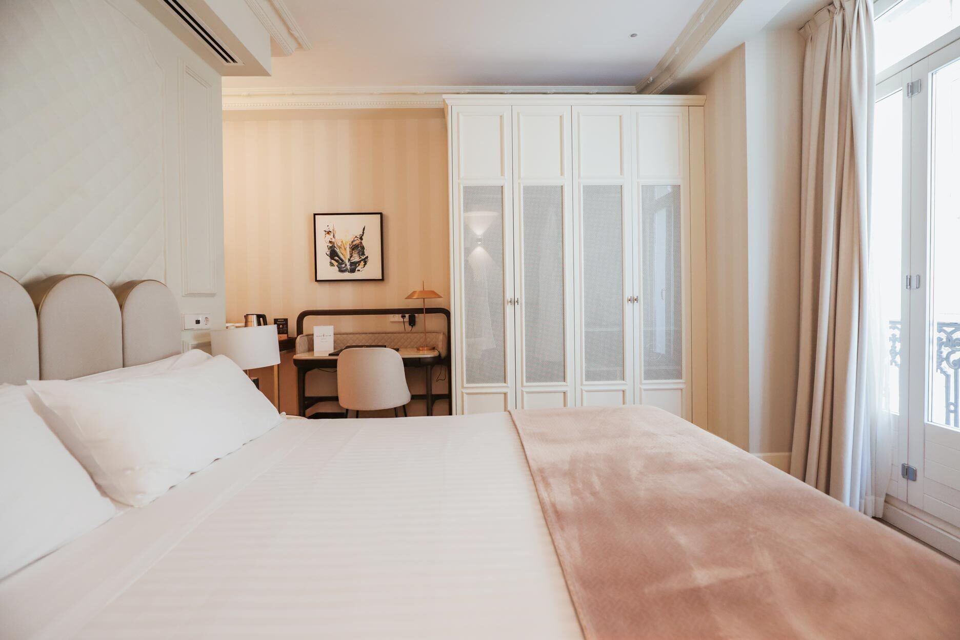 A hotel room with a large bed, a desk, a chair, a wardrobe, and a window with curtains.