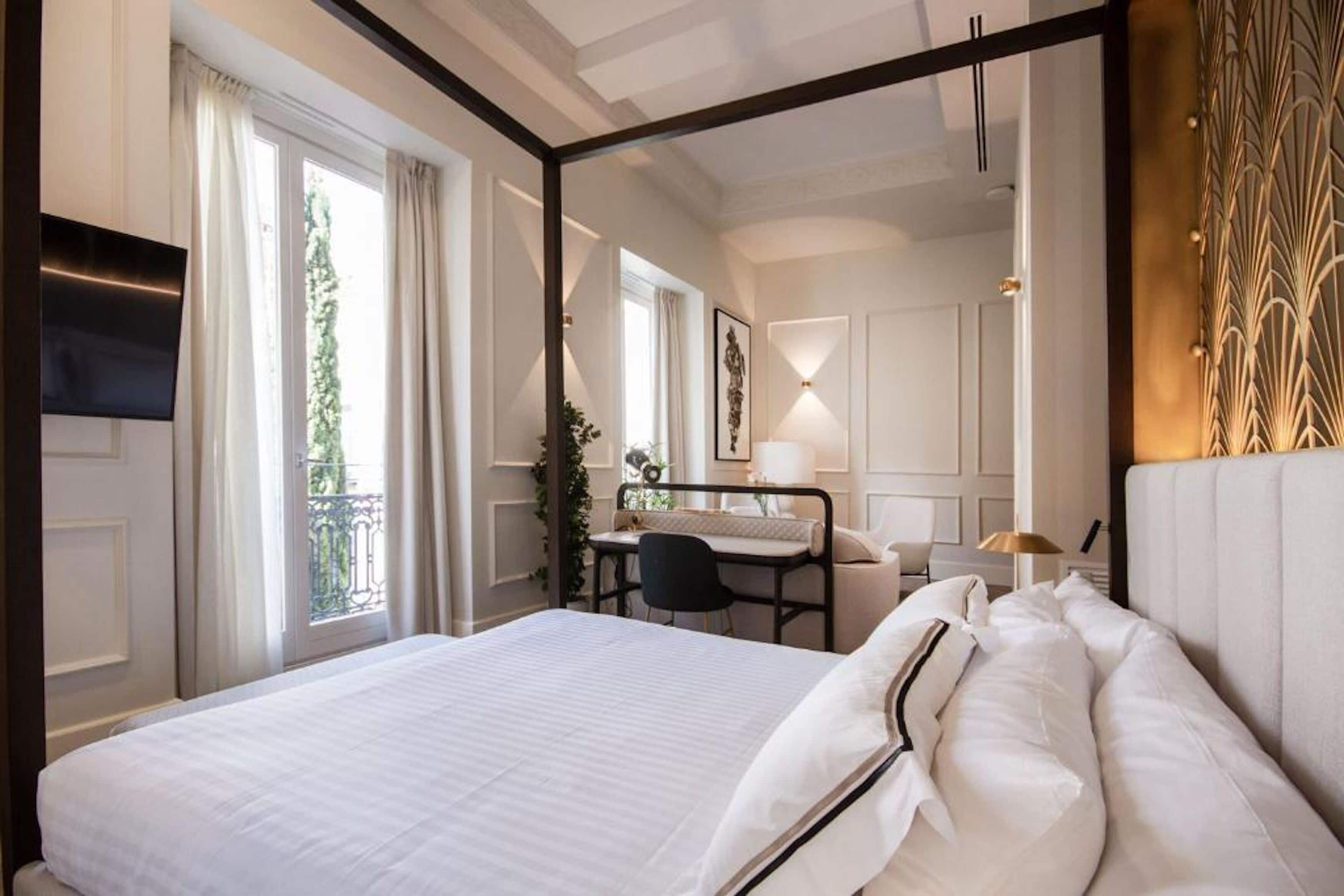 A hotel room with a large bed, a desk with a chair, a TV, and a balcony with curtains.