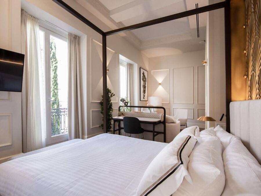 A hotel room with a large bed, a desk with a chair, a TV, and a balcony with curtains.