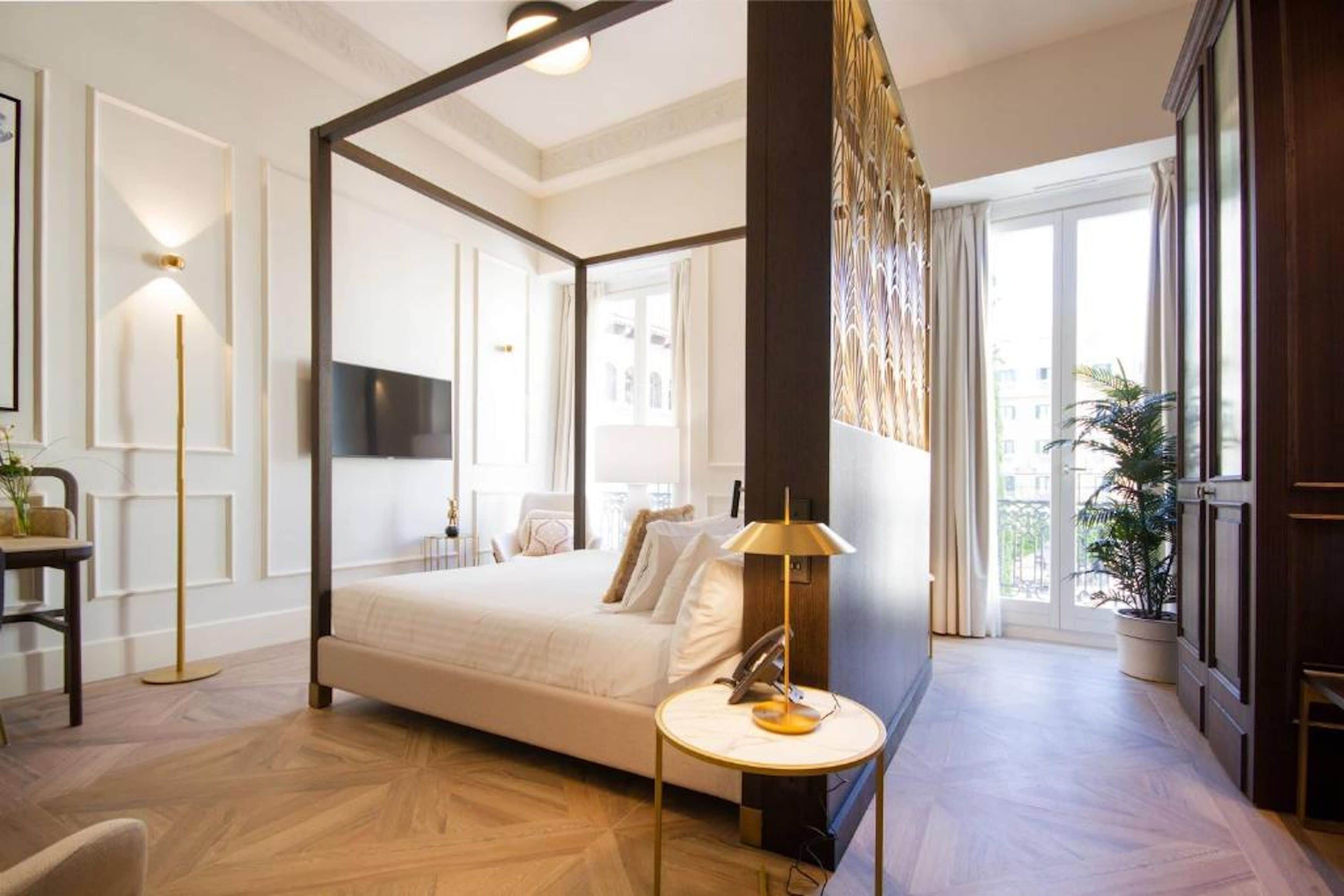 A four-poster bed in a well-lit room with a large window, a bedside table with a lamp, and a potted plant.