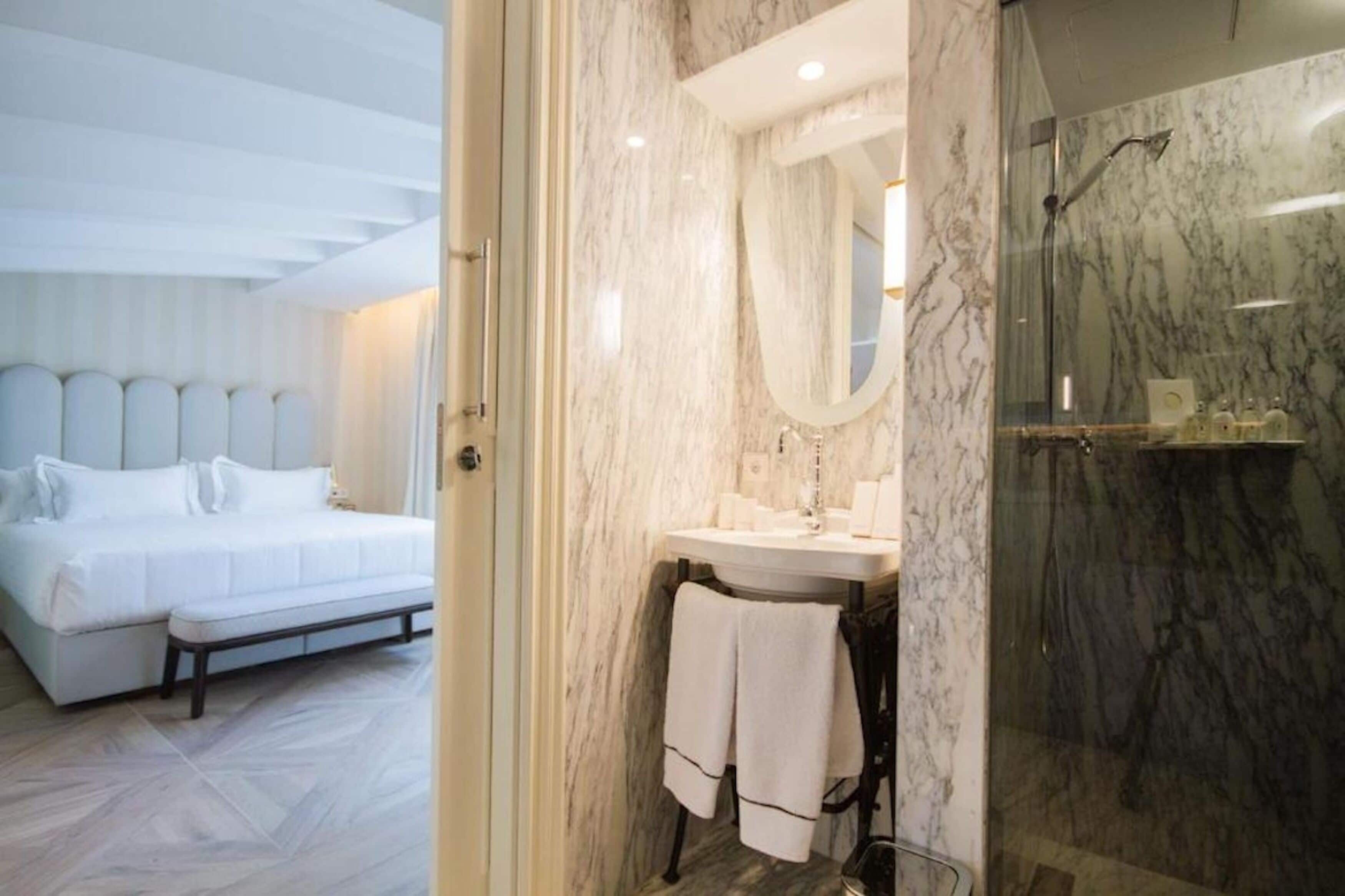 Un dormitorio moderno con una cama grande, un baño con lavamanos y ducha de mármol, y un pasillo con un espejo.