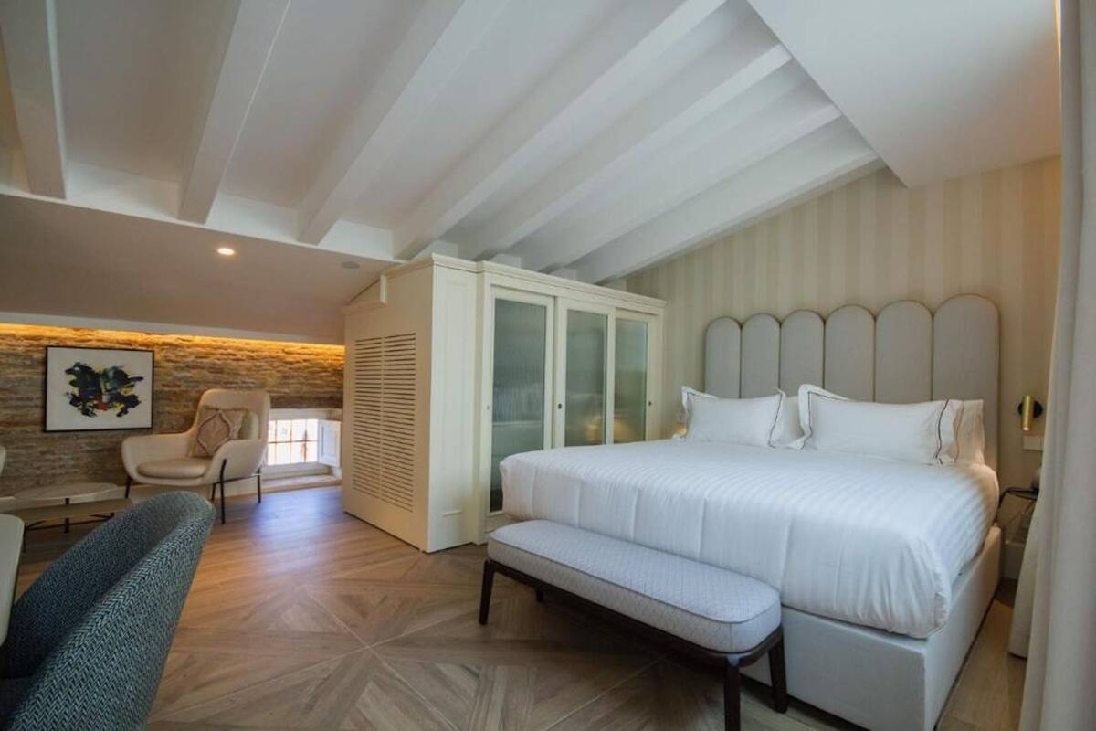 A bedroom with a large bed, a bench, a chair, a wardrobe, and a stone wall with artwork.