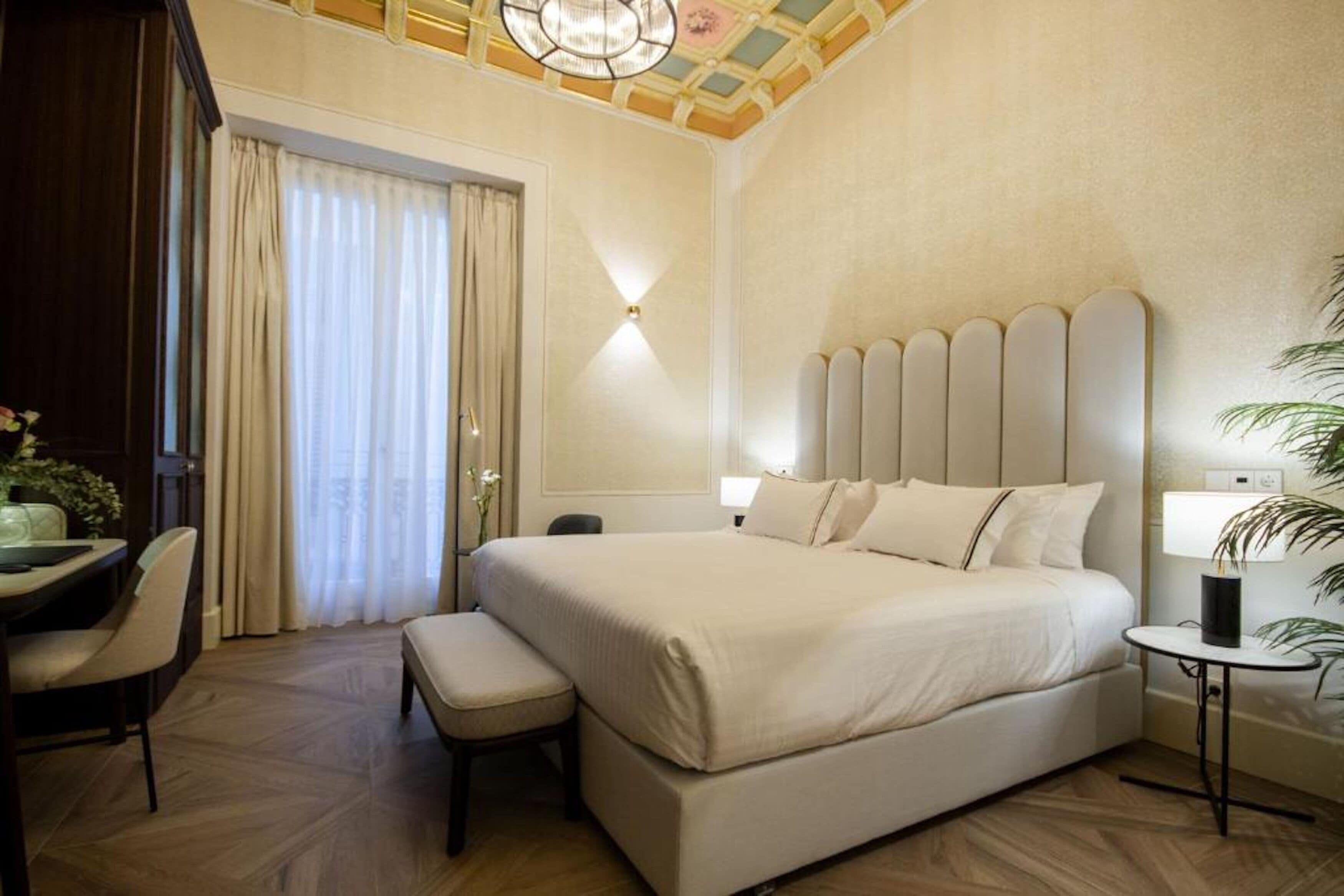 A hotel room with a large bed, a desk with a chair, a small table, and a potted plant.