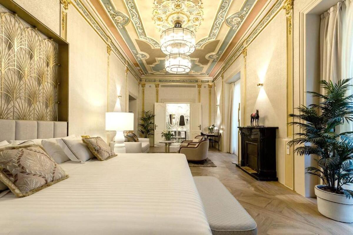 A luxurious hotel room with a large bed, a sitting area with a sofa and armchairs, a dining table, and a fireplace.