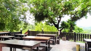 An outdoor seating area with wooden tables and benches, surrounded by a wooden fence and large trees.