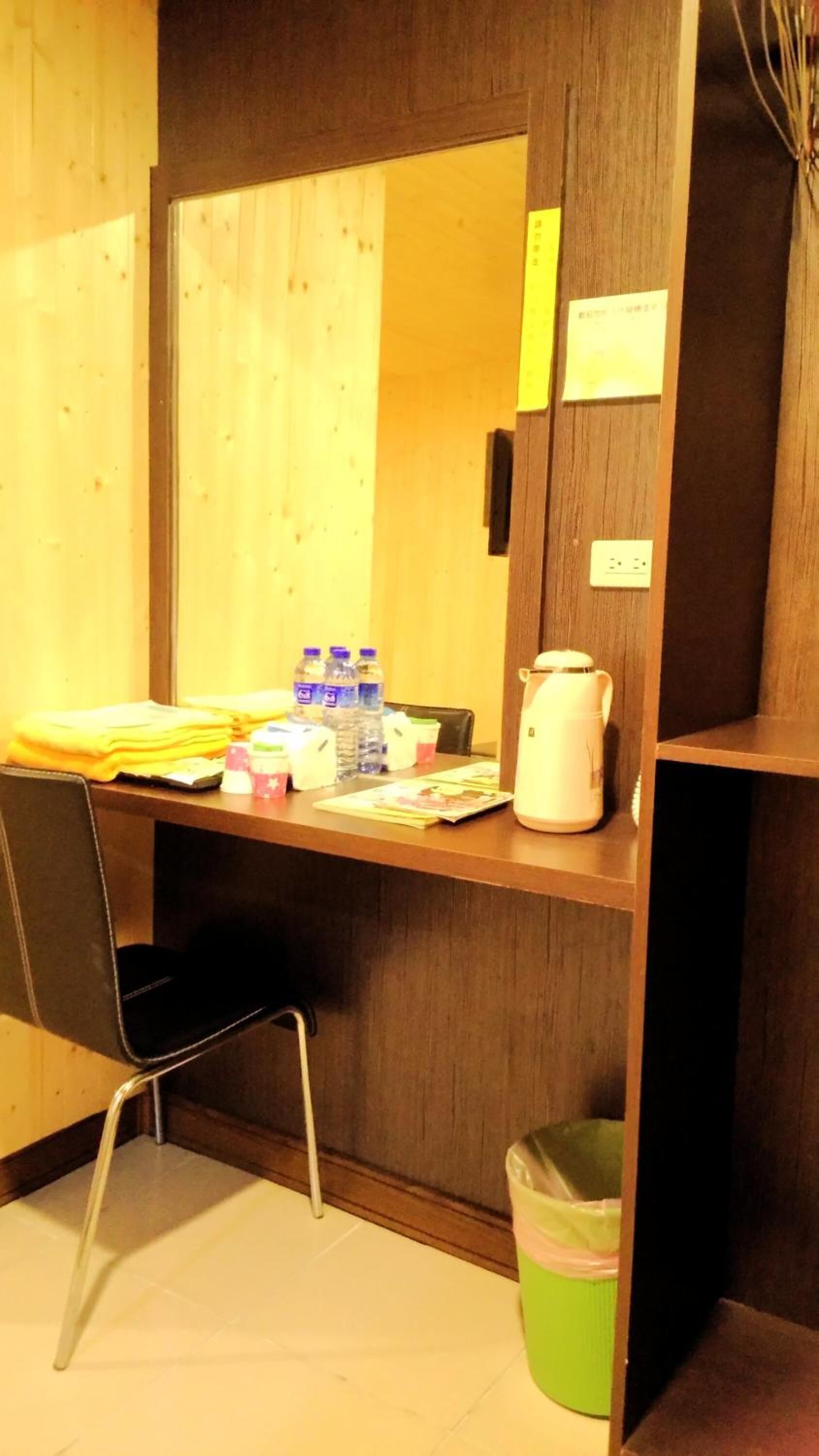 Una habitación de hotel pequeña con un escritorio de madera, una silla, un cubo de basura y una tetera.