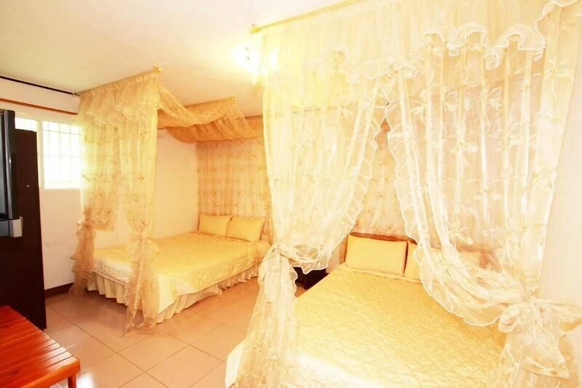 A room with two beds covered by sheer curtains, a wooden bench, and a television on the left wall.