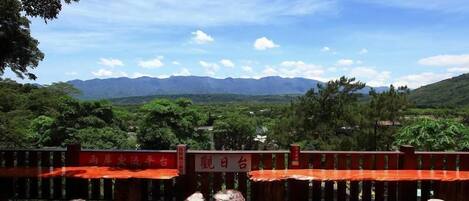 A terrace with wooden tables and chairs, surrounded by a wooden railing, overlooking a mountainous landscape.