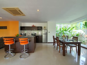 Una cocina moderna con una isla central, taburetes de bar, una mesa de comedor con sillas y vistas a exuberante vegetación a través de amplios ventanales.