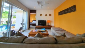 A modern living room with a sofa, a coffee table, a TV, and a laptop. There is a balcony with a pool view.