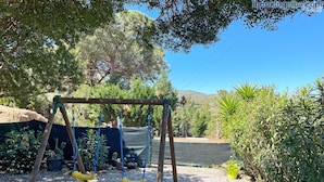 A wooden swing set with yellow seats in a garden area with gravel ground, surrounded by various plants and trees. - Appartamenti La Risacca - Appartamenti La Risacca - Trilo 4 VM (Capoliveri)