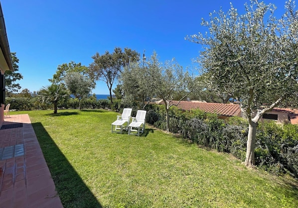 Un jardin bien entretenu, doté d’une terrasse, de deux fauteuils de jardin blancs et offrant une vue sur la mer.