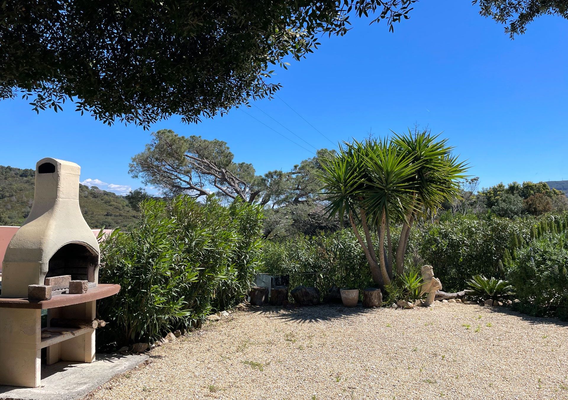 A rustic outdoor area with a stone oven, surrounded by gravel, trees, and a statue.