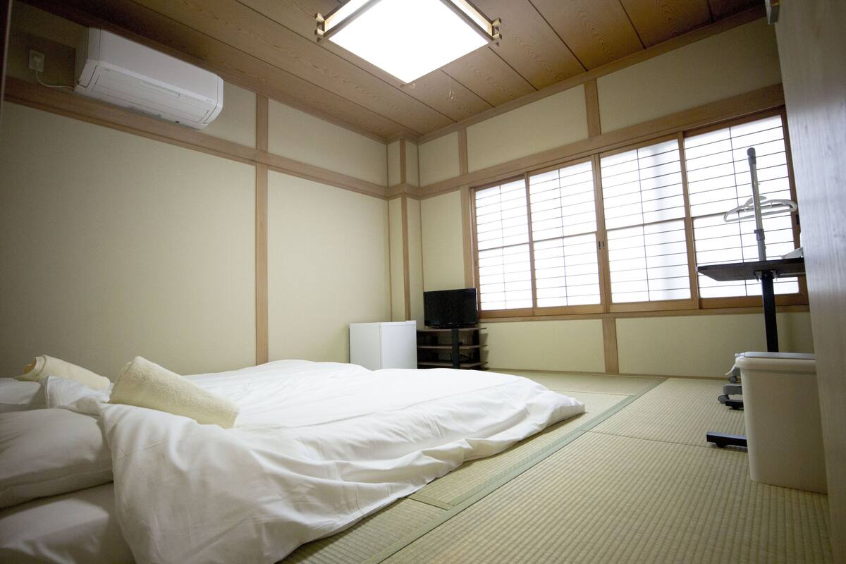 A traditional Japanese-style room with tatami flooring, a bed with white bedding, a television, and a heater.