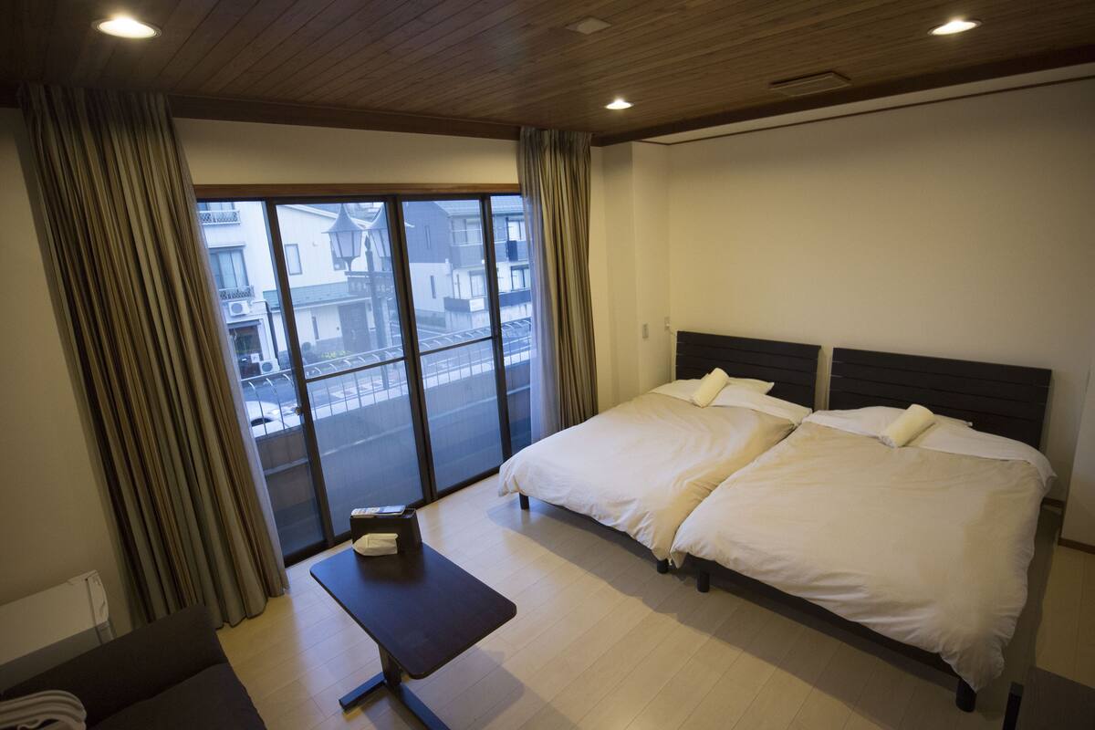 A hotel room with a large bed, a small table, and a view of the city through a large window.