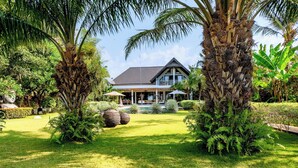 Une grande maison avec un toit sombre, entourée d’une végétation luxuriante et de palmiers.