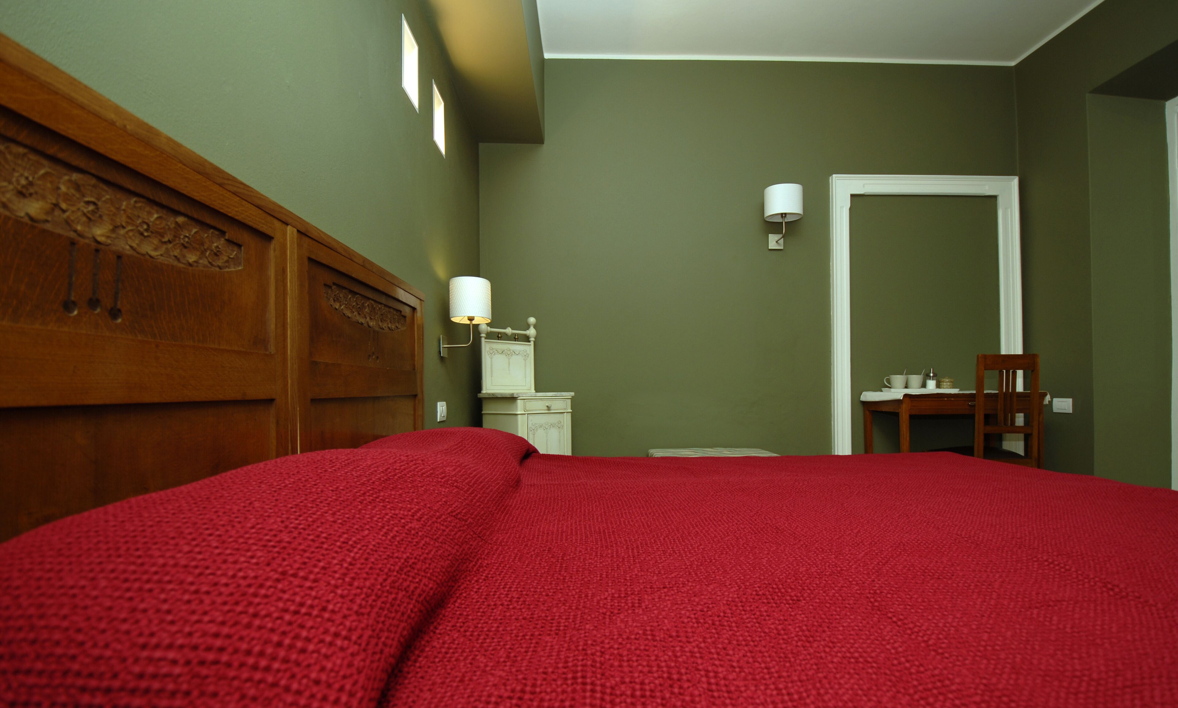 A bedroom with a wooden headboard, a red bedspread, a nightstand with a lamp, a small table with a chair, and a door leading to another room.