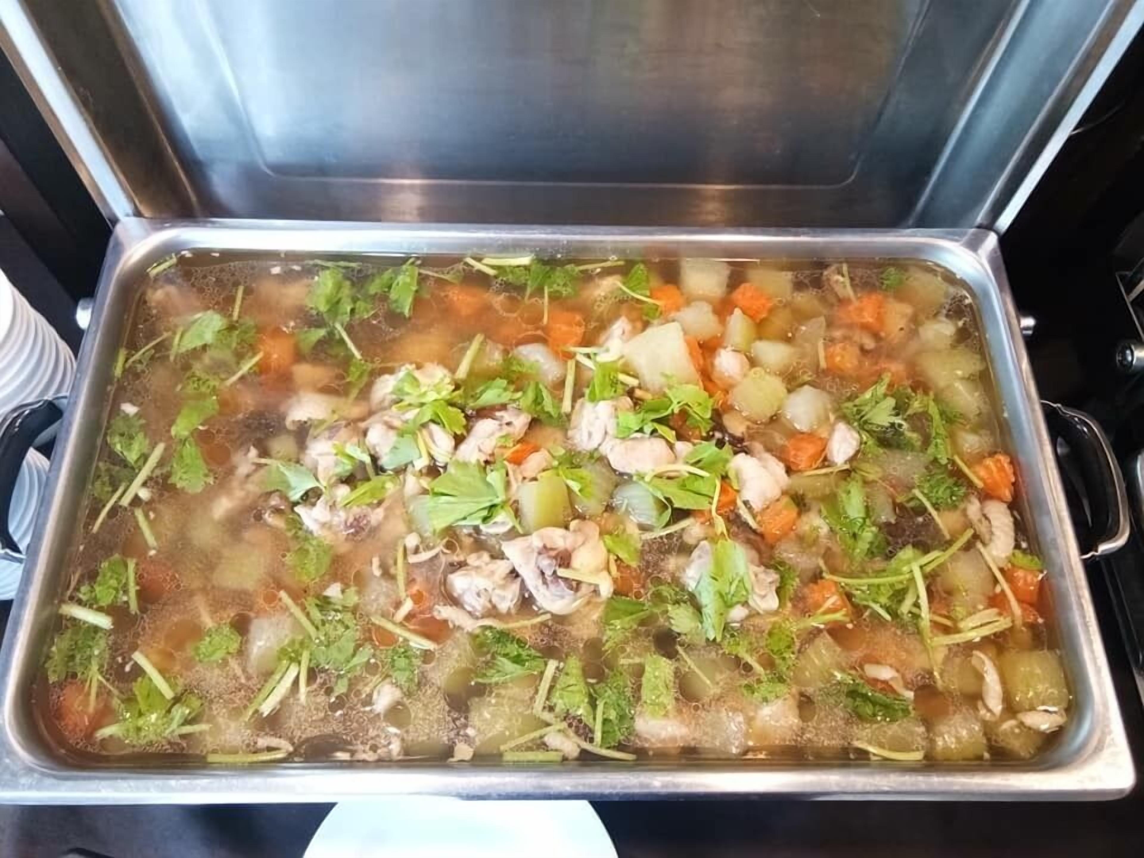 Una olla de acero inoxidable grande llena de un guiso con carne, verduras y hierbas aromáticas.