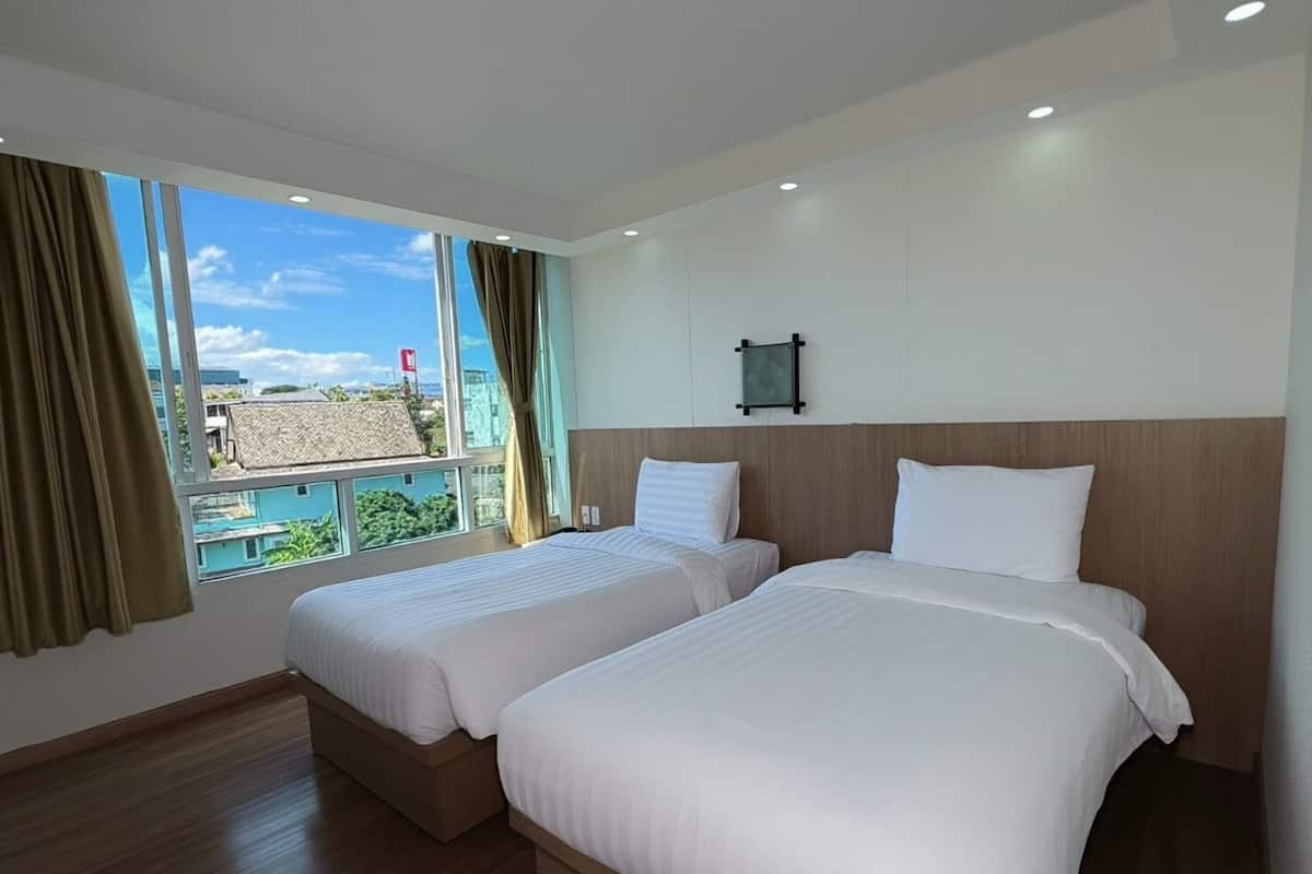 A hotel room with two beds, a large window with curtains, a view of buildings, and a wall-mounted light fixture.