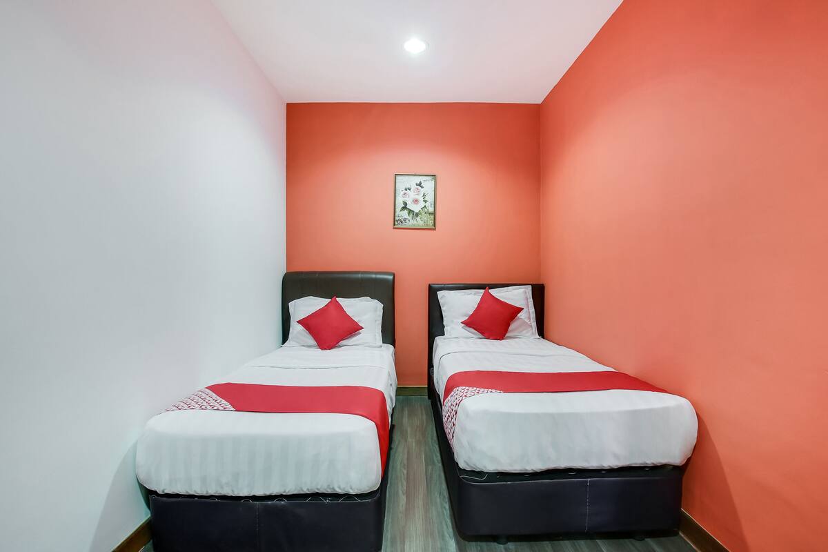 Ein Zimmer mit zwei Einzelbetten, jeweils mit rotem und weißem Bettbezug und passenden Kissen. Die Wände sind in einem warmen Orange-Ton gestrichen, und über den Betten hängt ein gerahmtes Bild.