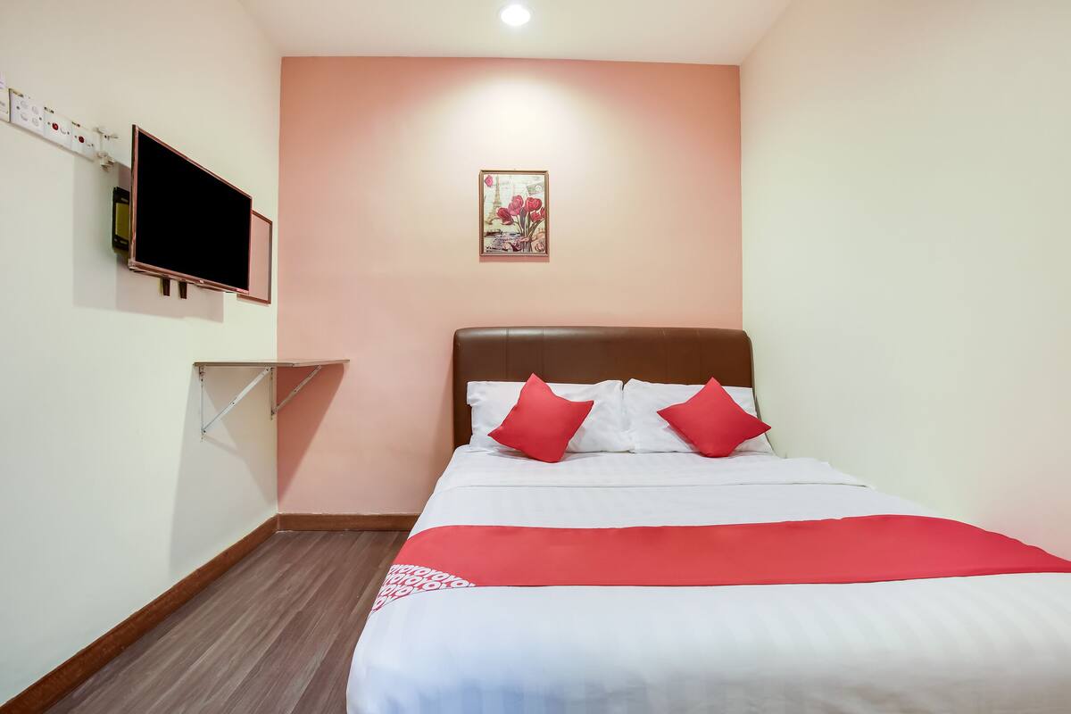 Ein ordentlich bezogenes Bett mit roter und weißer Bettwäsche, ein an der Wand befestigter Flachbildfernseher und eine Wandleuchte.
